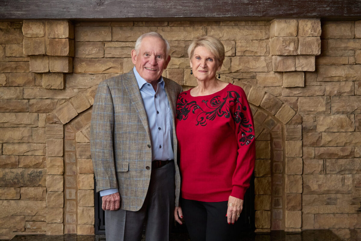Two people pose for a portrait in front of a stone fireplace, with one wearing a plaid sport coat and the other a red sweater with black detailing.