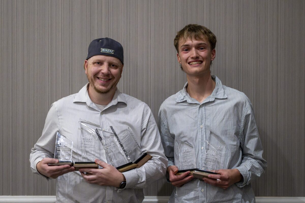 Two award recipients stand indoors holding multiple glass plaques and trophies while posing for a photo against a neutral wall.