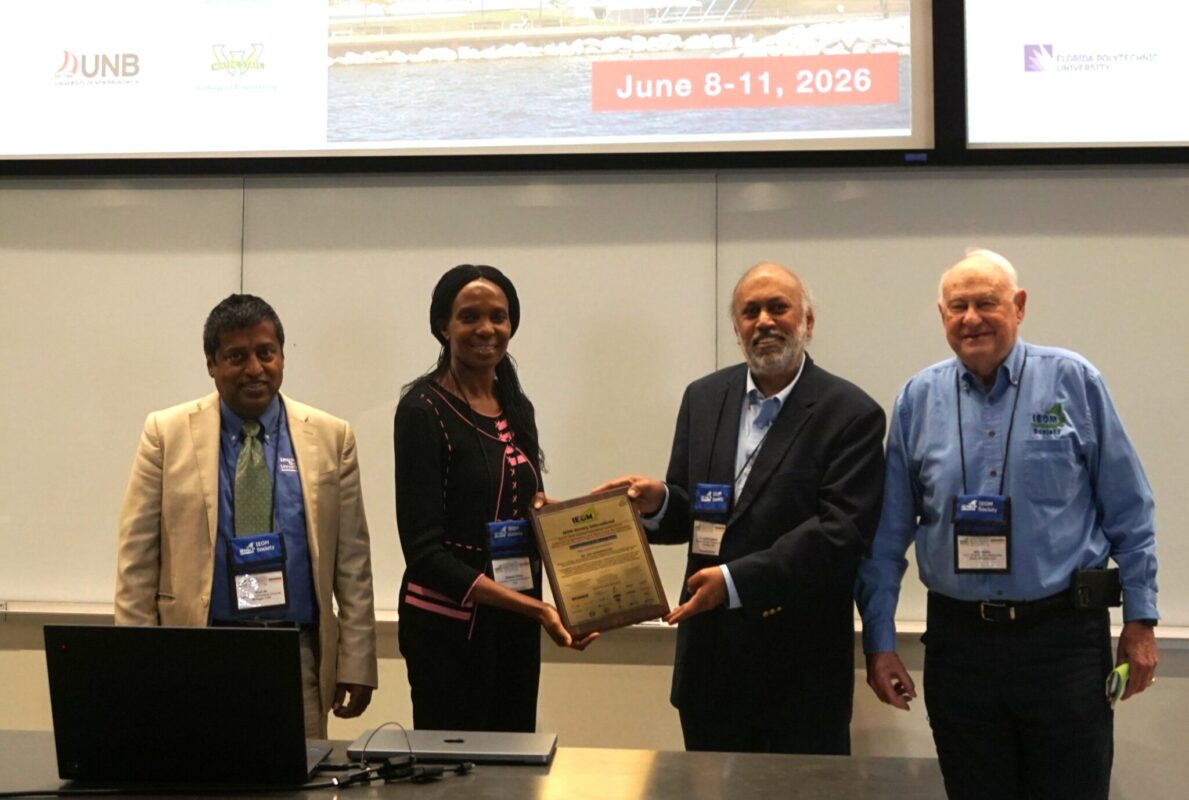 Four conference attendees stand at the front of a meeting room while two individuals present and receive a plaque. Presentation screens and conference signage are visible in the background.