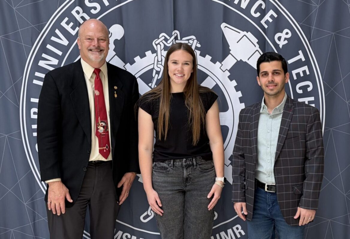 Three people pose in front of a Missouri University of Science and Technology backdrop featuring a gear design and university name. Two people wear business attire, and one wears a black shirt and gray pants.
