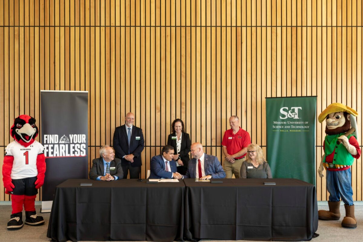 People gathered around a table during a signing ceremony, with SEMO and Missouri S&T banners in the background and mascots standing on either side.