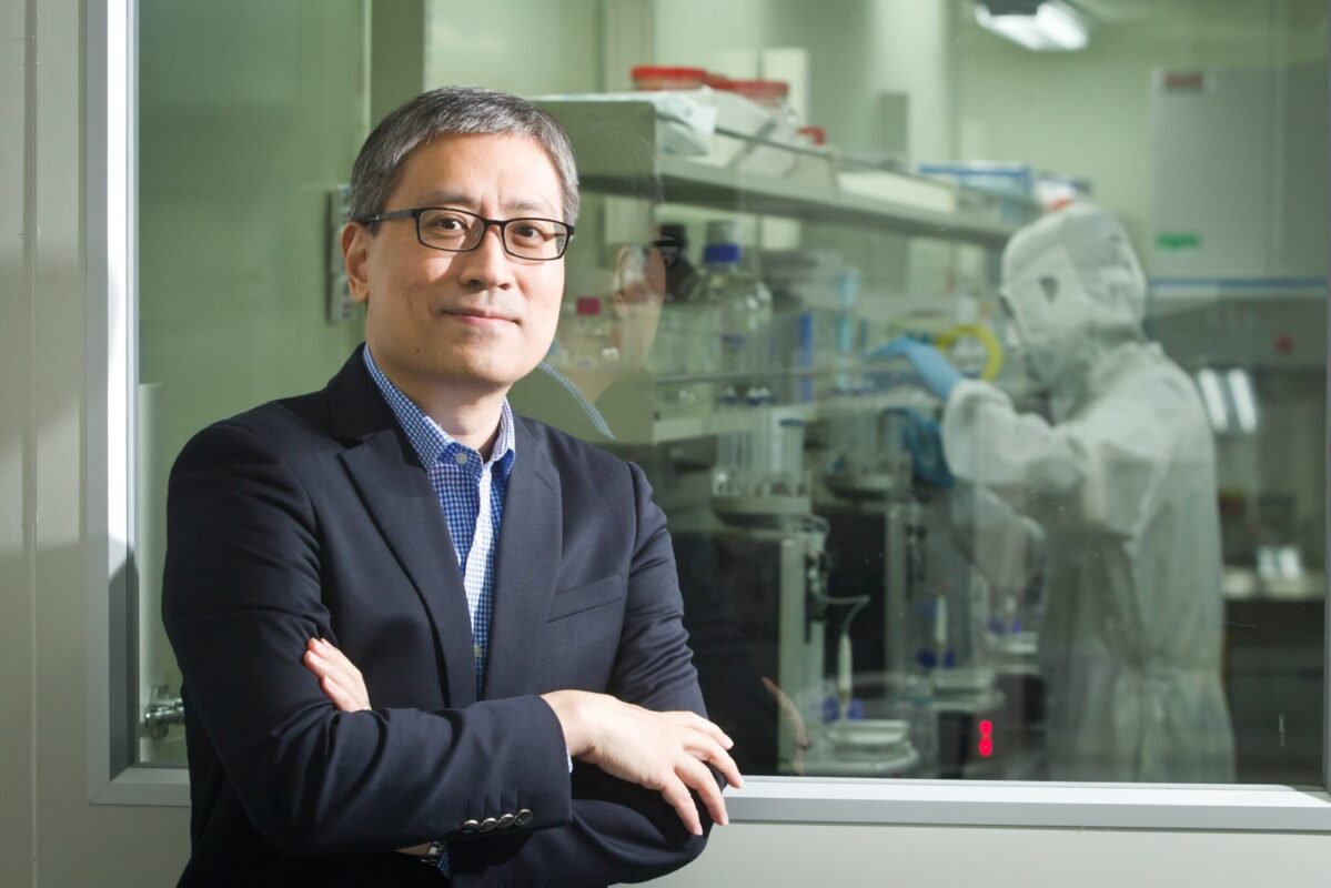 Dr. Huang in a dark blazer and glasses standing with arms crossed beside a laboratory observation window. In the background, a researcher wearing full protective cleanroom gear works with equipment and containers inside a laboratory. The image highlights a research and scientific innovation environment.