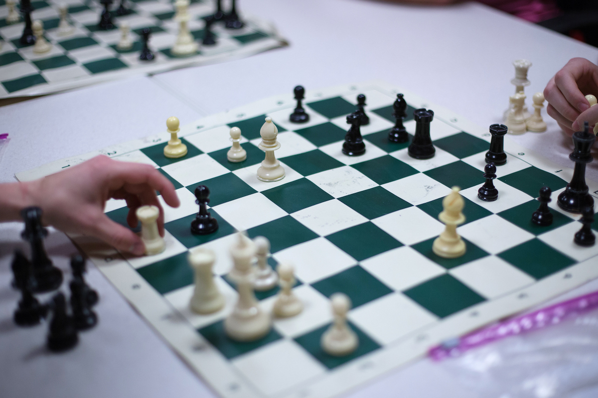 A chessboard sits in the foreground in focus as students play on other tables in the background.