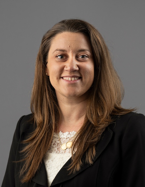 Professional headshot of a person with long hair wearing a dark blazer over a white patterned blouse, photographed against a plain gray background.