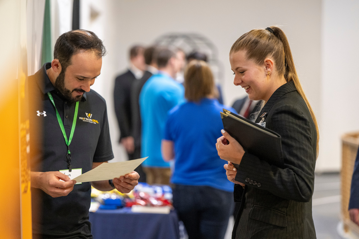 Missouri S&T student presents a resume to a prospective employer at a campus career fair, with other recruiters and attendees networking in the background.