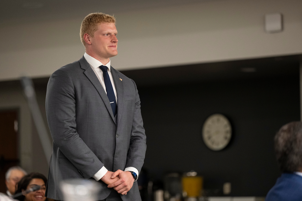 Owen Fraser stands in a meeting wearing a gray suit, white dress shirt and dark tie.
