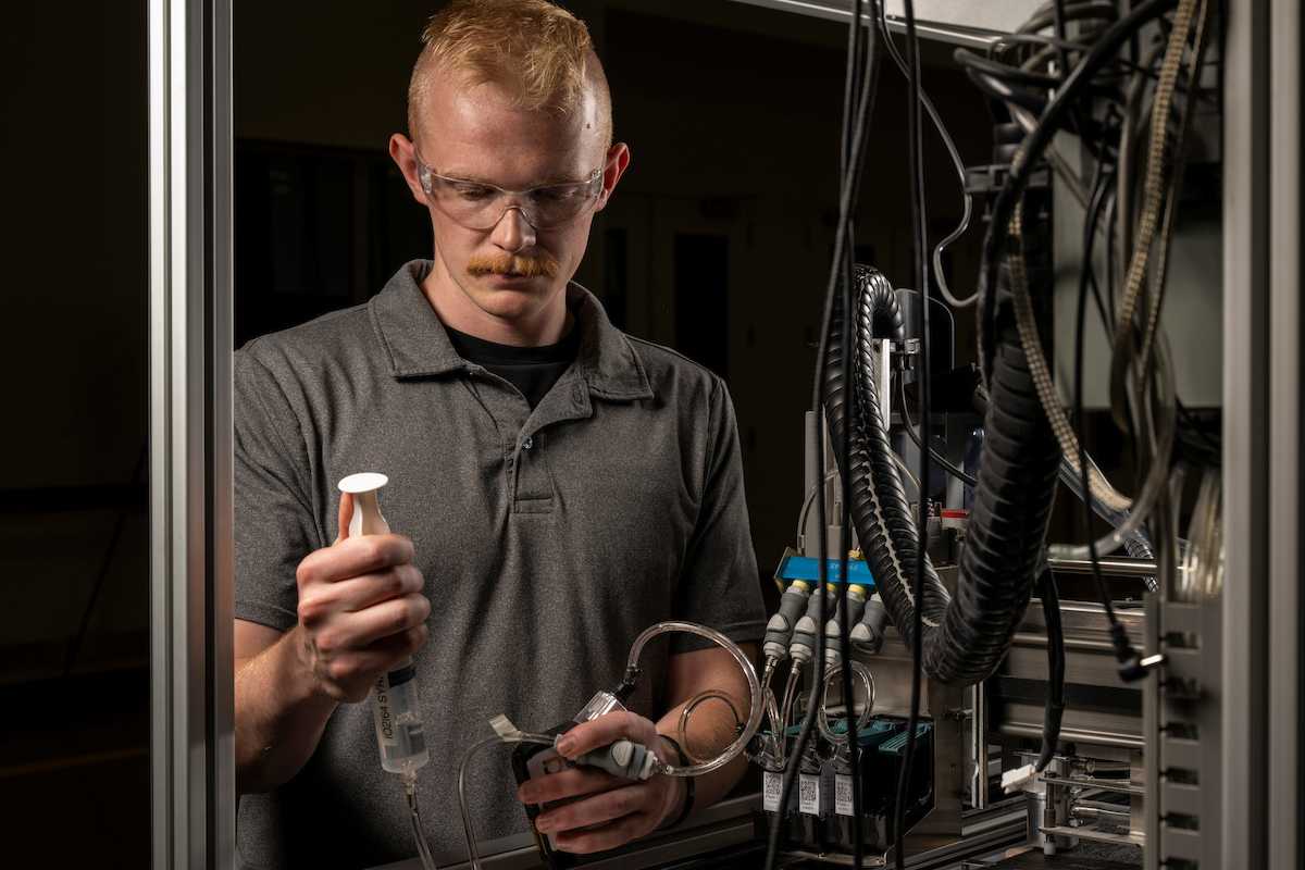 Person wearing safety glasses operates laboratory equipment, using a syringe-like device connected to tubing within an industrial research or testing setup.