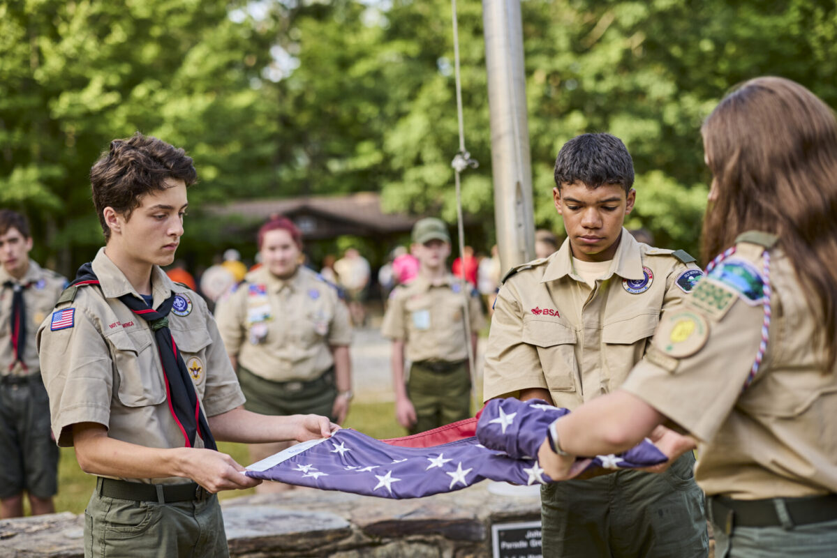Three Scouts in uniform carefully fold an American flag during a flag ceremony outdoors. Additional Scouts stand nearby in the background, with trees and a campground shelter visible behind them.