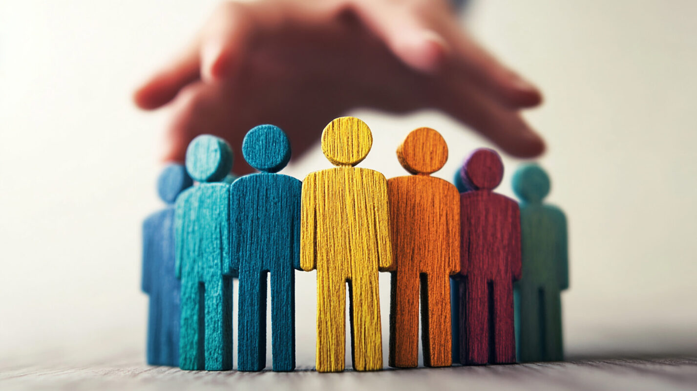 Colorful wooden figures stand together beneath protective hands, symbolizing community, support and inclusion.