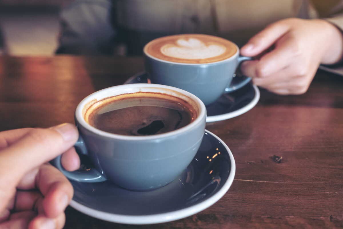 Two cups of coffee on a wooden table, with one person holding a black coffee in the foreground and another person holding a latte with foam art in the background. The image is softly lit and focused on the coffee cups.