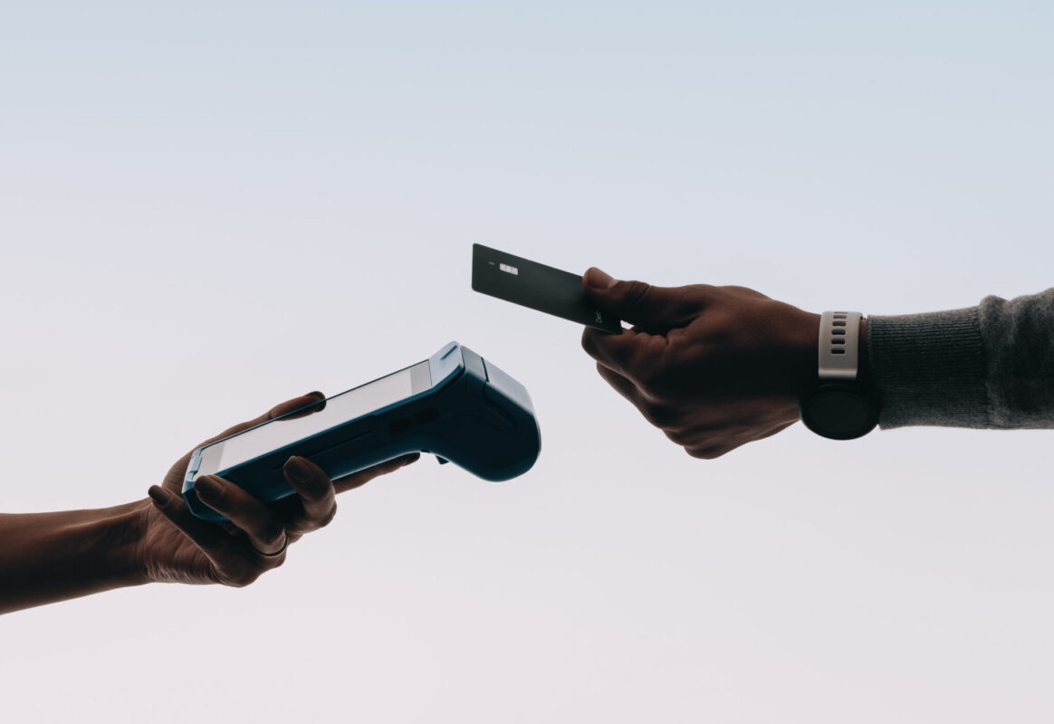 Two hands exchange a card and a payment terminal against a light background, illustrating a contactless transaction or payment process.