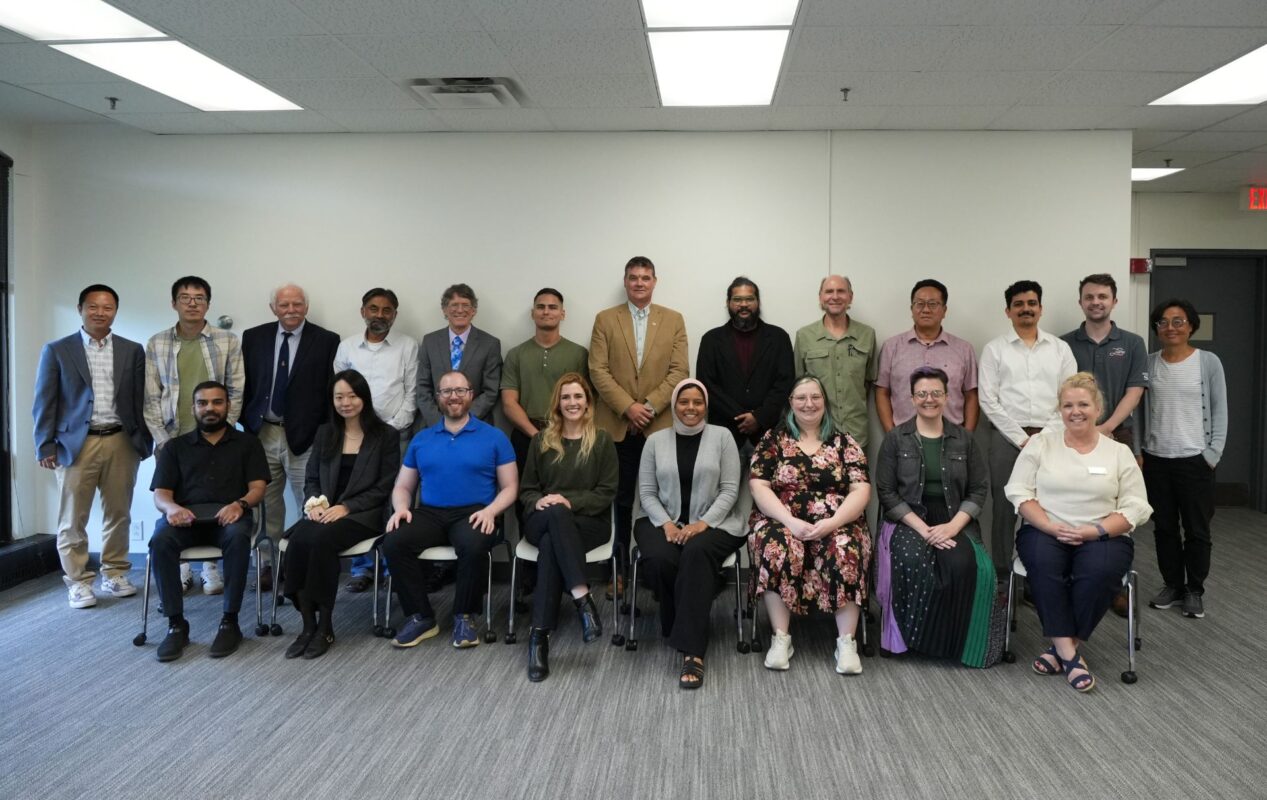 Group portrait of approximately 20 new faculty arranged in two rows, with seated participants in the front row and standing participants behind them. The group poses for a photo against a plain white wall, with overhead lights and gray carpeting visible.