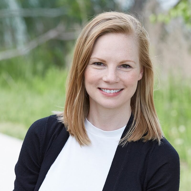 Professional headshot of a person with shoulder-length light hair, wearing a white top and dark cardigan, facing the camera outdoors. The background is softly blurred with green grass, trees, and a pathway visible in natural daylight.
