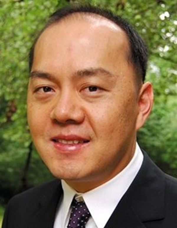 Professional headshot of a person wearing a dark suit, white shirt and patterned tie, photographed outdoors with green foliage softly blurred in the background.