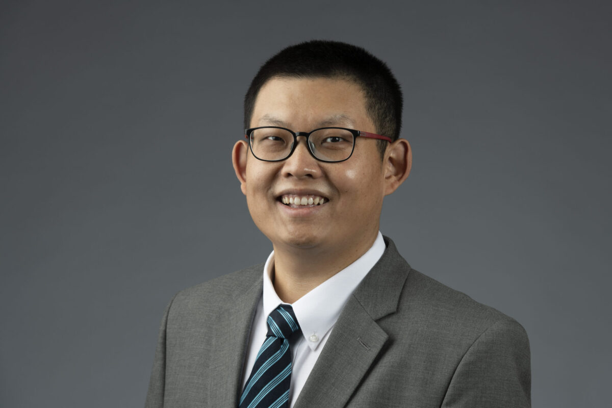 Professional headshot of a person wearing glasses, a gray suit jacket and a blue striped tie against a gray background.