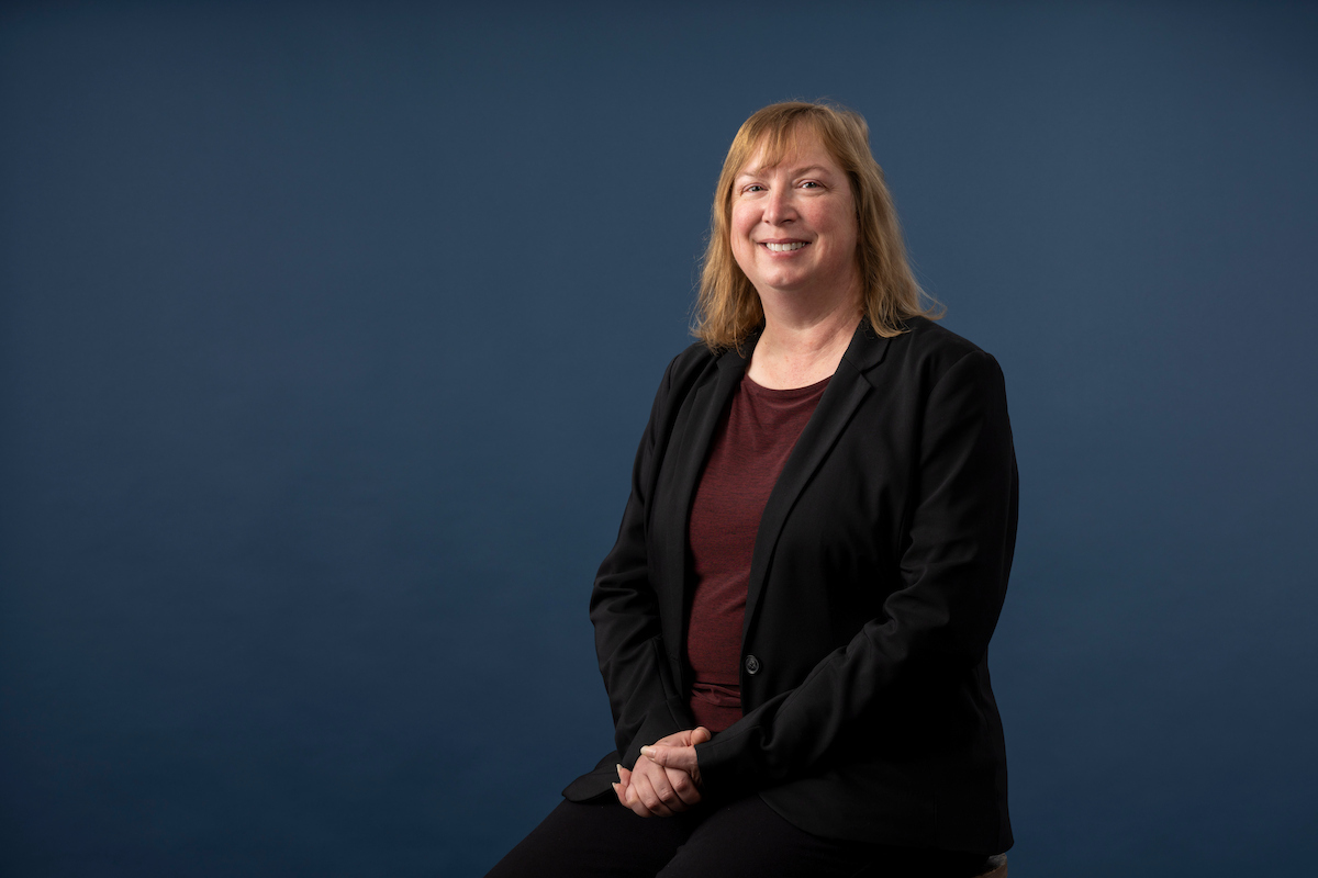 Professional portrait of a person seated against a blue background, wearing a dark blazer and maroon top.