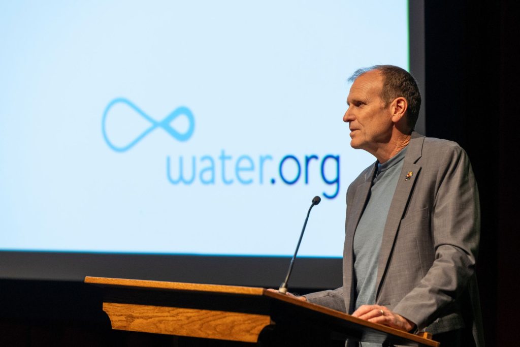 A man wearing a brown sport coat over a grey T-shirt gives a talk behind a podium with the "water.org" logo projected on the screen behind him.