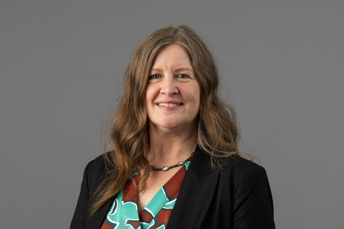 Professional headshot of a person wearing a dark blazer and patterned blouse against a gray background.