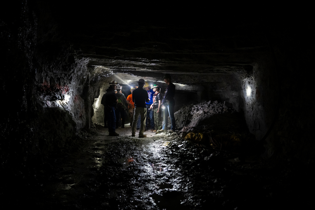 A group of participants wearing safety gear stand inside Missouri S&T's Experimental Mine. Lights illuminate the underground tunnel, rocky walls and damp mine floor.