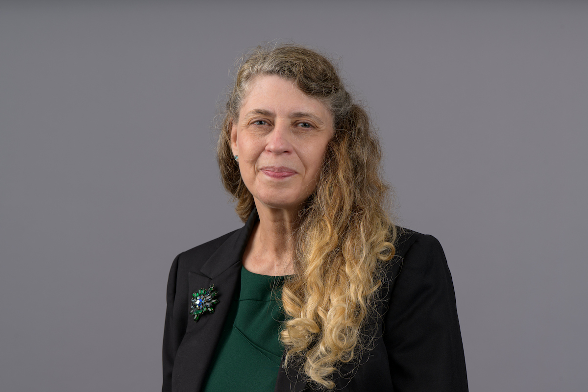 Professional headshot of a person wearing a dark blazer and green top against a gray background.