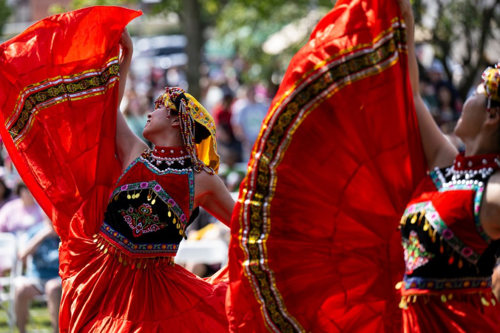 Performers in colorful embroidered costumes hold flowing red fabric overhead while dancing outdoors before a crowd.
