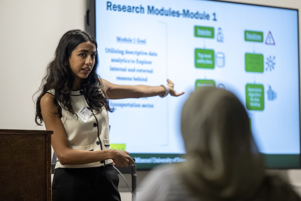 A presenter gestures toward a screen displaying a research model during a presentation.