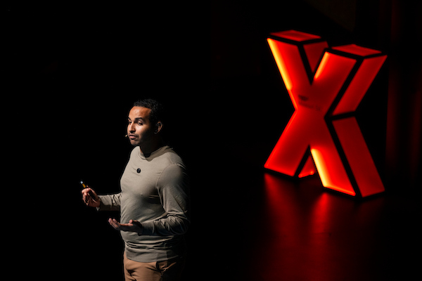 A speaker stands on a dark stage holding a presentation remote and gesturing, with a large illuminated red X behind the speaker.