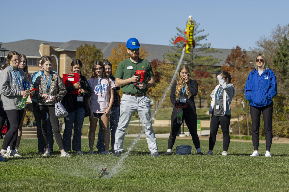A group watches an outdoor demonstration as a person in a blue hard hat operates equipment spraying water across a grassy field.
