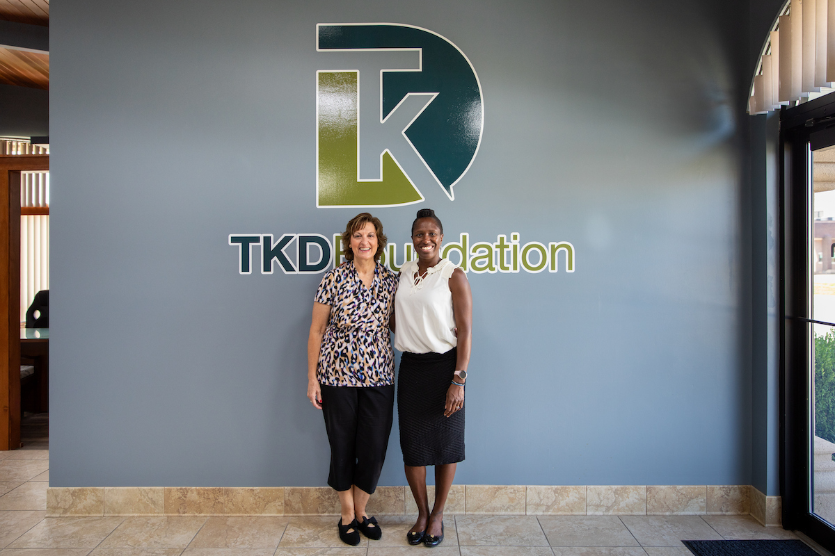 Two people stand in front of a wall displaying the TKD Foundation logo and name inside an office building.