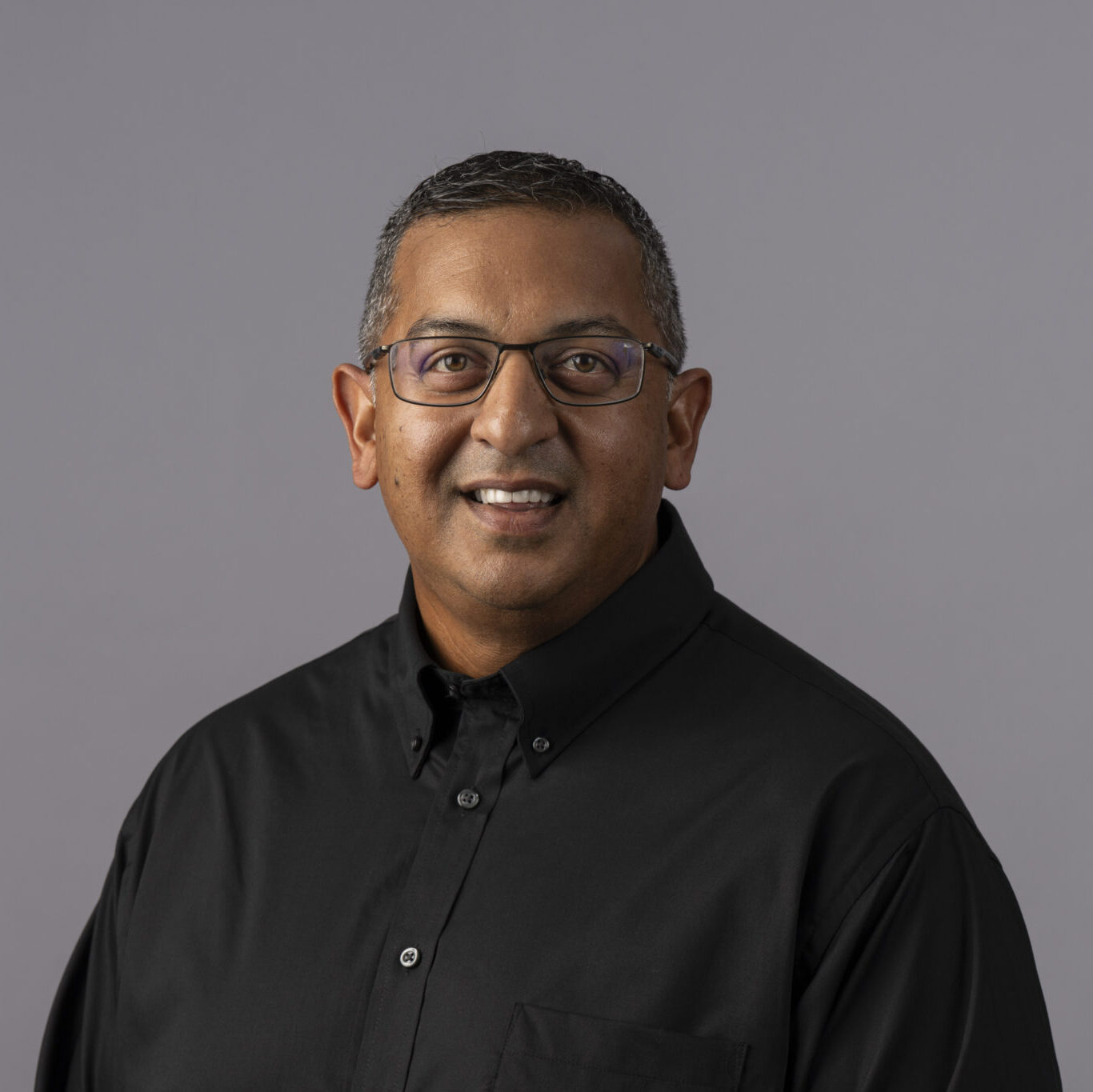 A person with short hair and glasses smiles while wearing a black button-up shirt against a plain gray background.