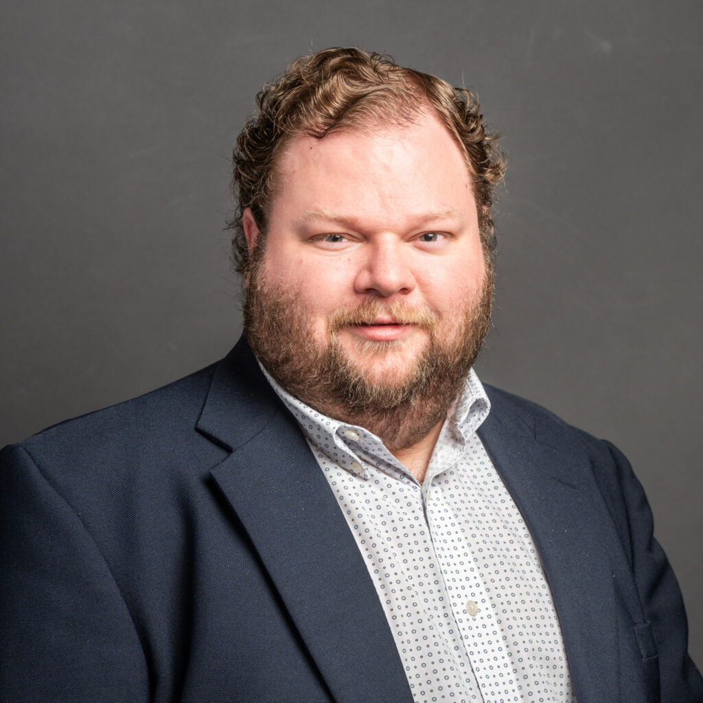 Professional headshot of a person wearing a dark blue blazer over a light-colored patterned button-down shirt, facing the camera against a plain dark gray background.