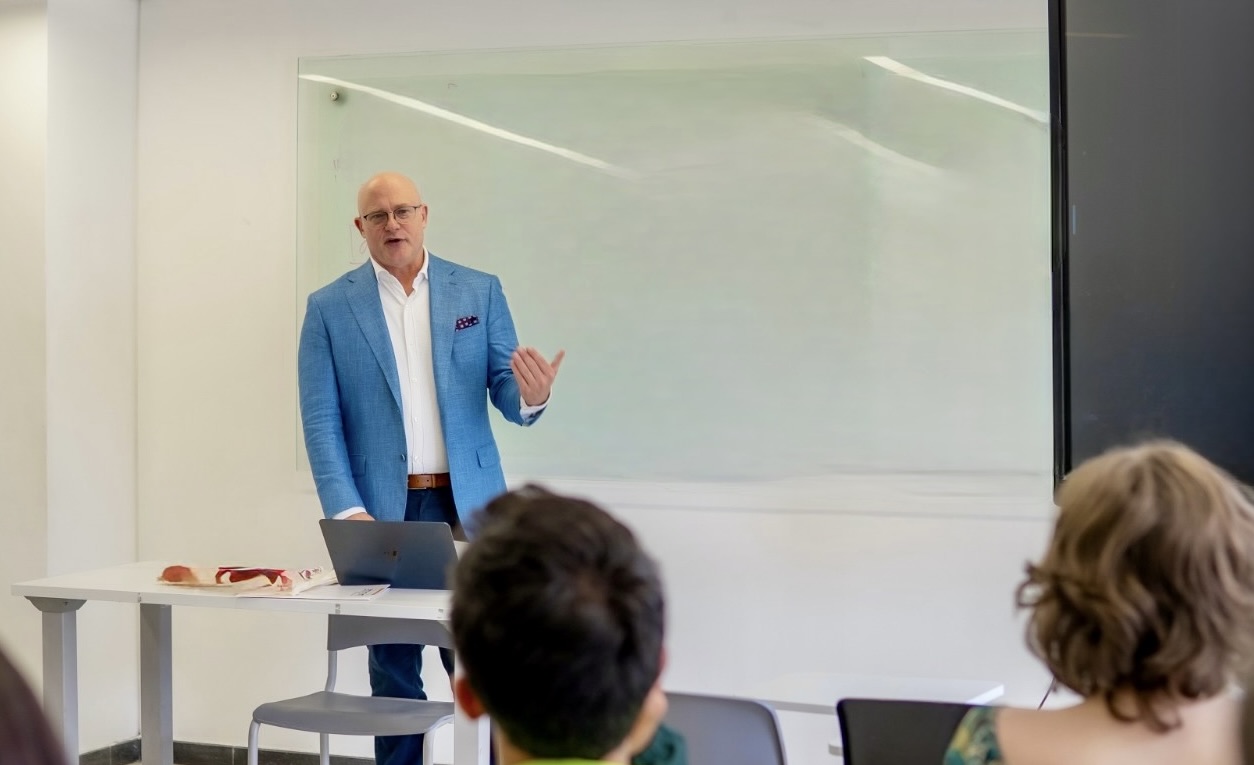 A man wearing a light blue blazer and white button up shirt teaches a class.