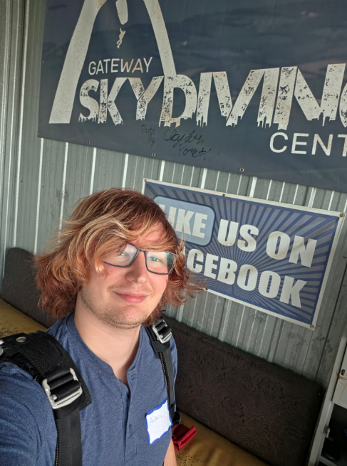 A person wearing glasses and a backpack takes a selfie inside the Gateway Skydiving Center, with signs for the skydiving center and its Facebook page visible in the background.