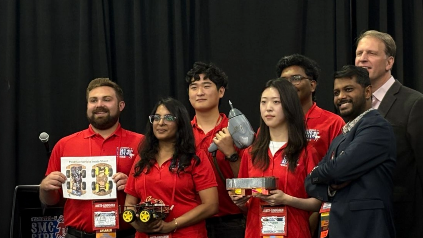 Group of six conference participants wearing red event shirts stand on a stage holding awards and display items while posing for a photo. Two additional individuals stand behind and beside the group. A black curtain forms the background.