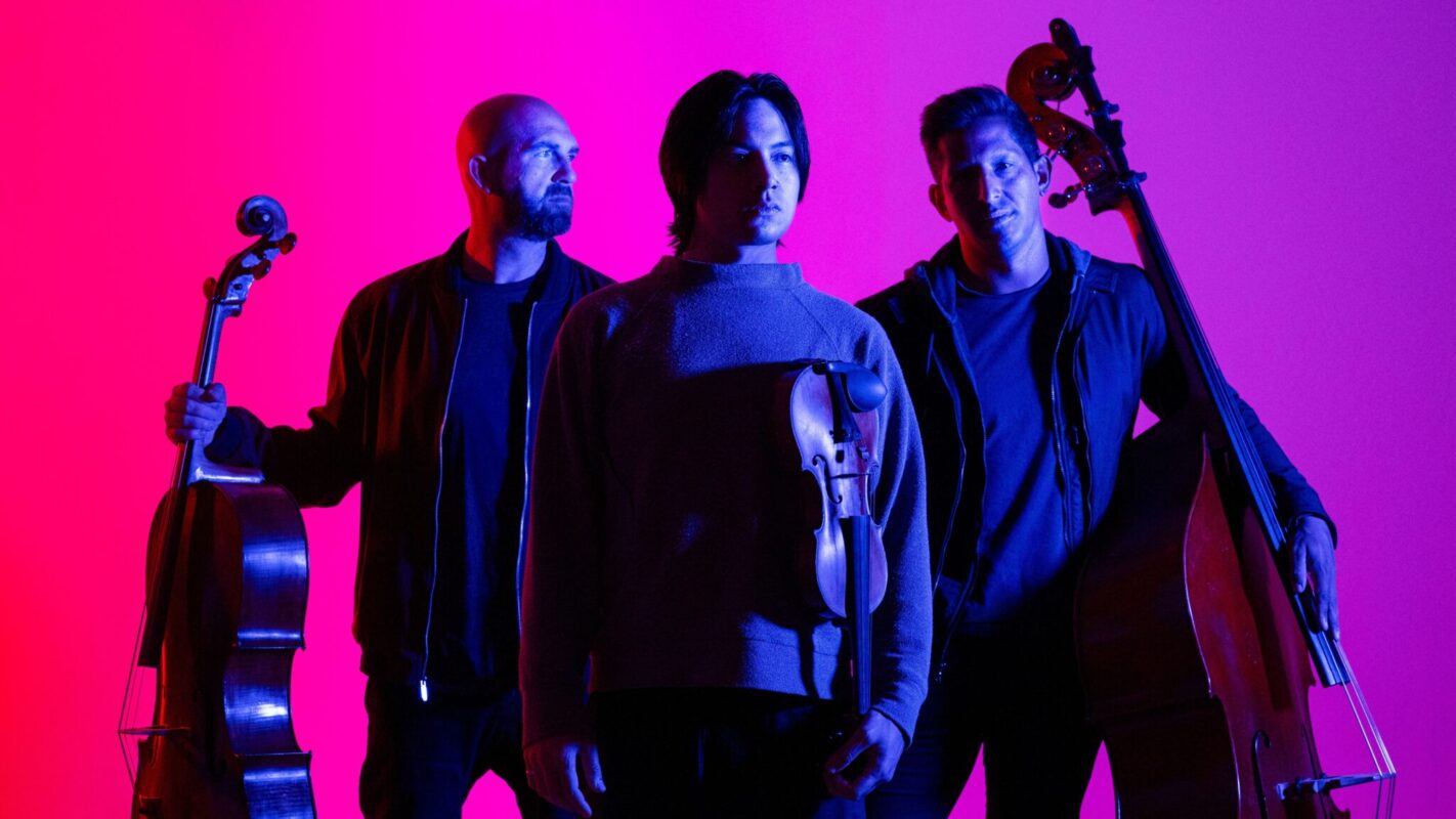 Promotional image of the string trio Simply Three standing with their instruments against a vibrant pink and purple gradient background. One musician stands in the center holding a violin, flanked by two musicians holding upright string instruments.