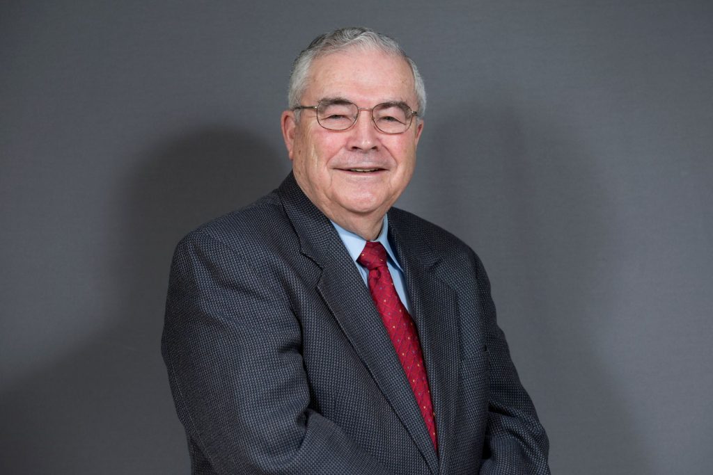Portrait of a person wearing glasses, a dark patterned blazer, white shirt and red tie, seated against a plain gray background.