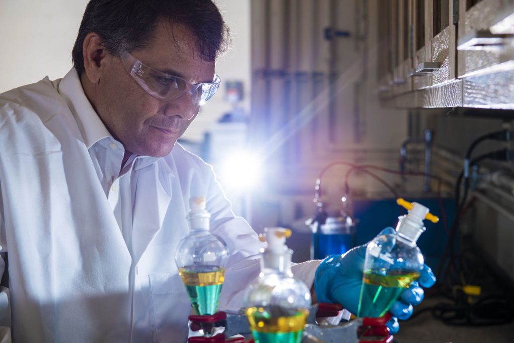 Researcher wearing safety glasses, a white lab coat and blue gloves examines laboratory glassware containing bright green and yellow liquids on a lab bench.