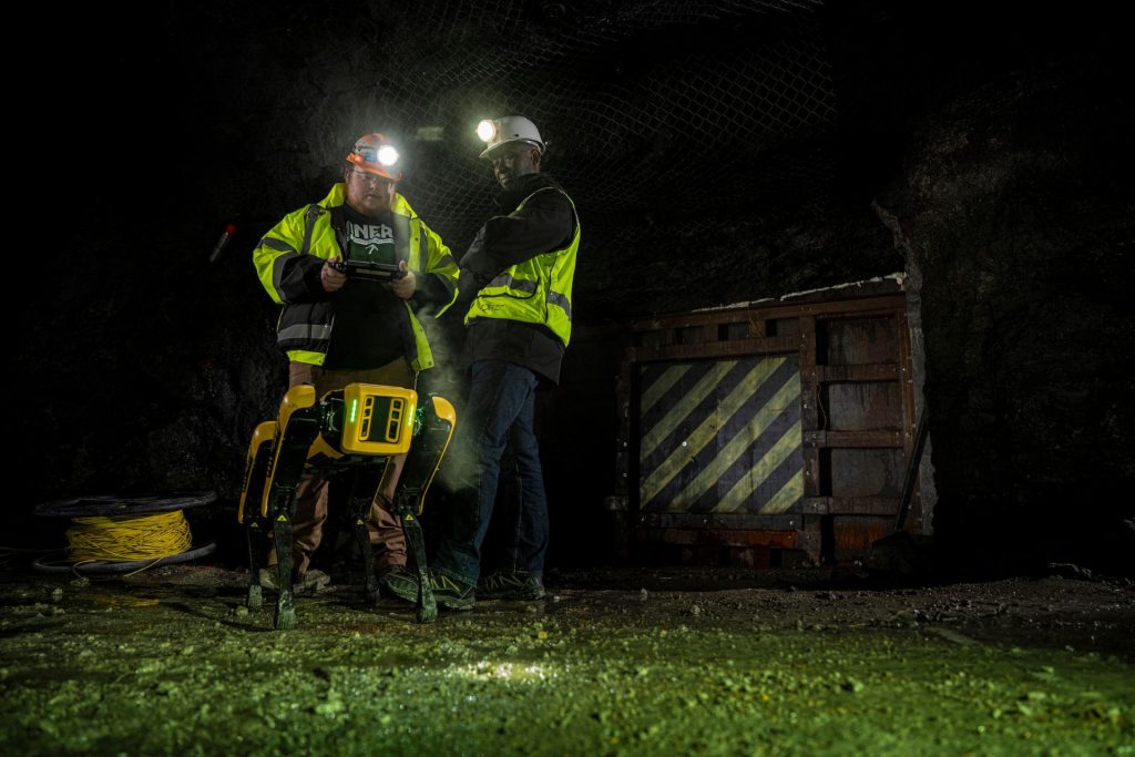 Two workers wearing high-visibility jackets and headlamps operate drilling equipment at night beside a large industrial container.