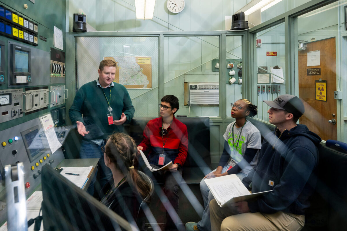 Five people gather around a large control panel in a glass-walled laboratory control room.