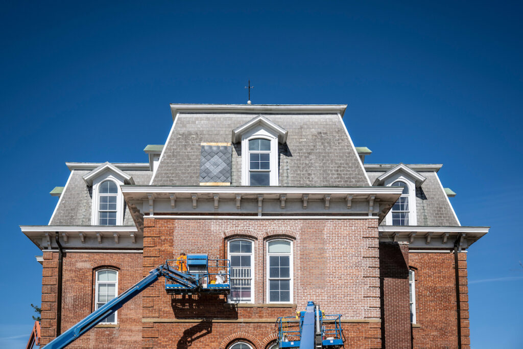 Front view of a historic red-brick building with a steep gray mansard roof, dormer windows and decorative architectural details under a clear blue sky. Blue construction lifts stand in front of the building.