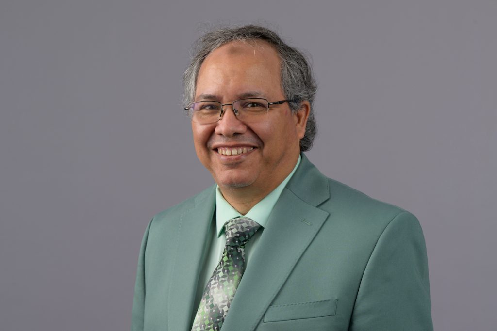 Portrait of a person wearing glasses, a light green suit jacket, white shirt and patterned green tie against a plain gray background.