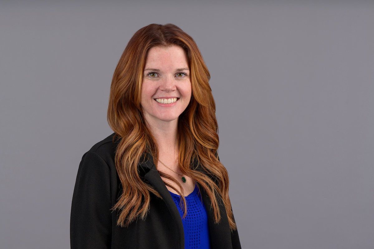 Portrait of a person with long red hair wearing a black jacket over a blue top, posed against a plain gray background.
