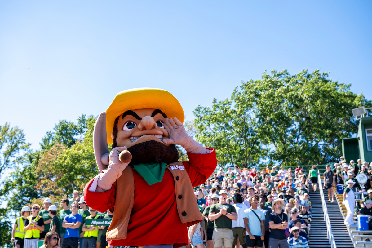 Joe Miner stands in front of a large crowd seated on outdoor bleachers. The mascot holds one hand near its mouth as if calling out to the audience. Trees and a clear blue sky are visible in the background.