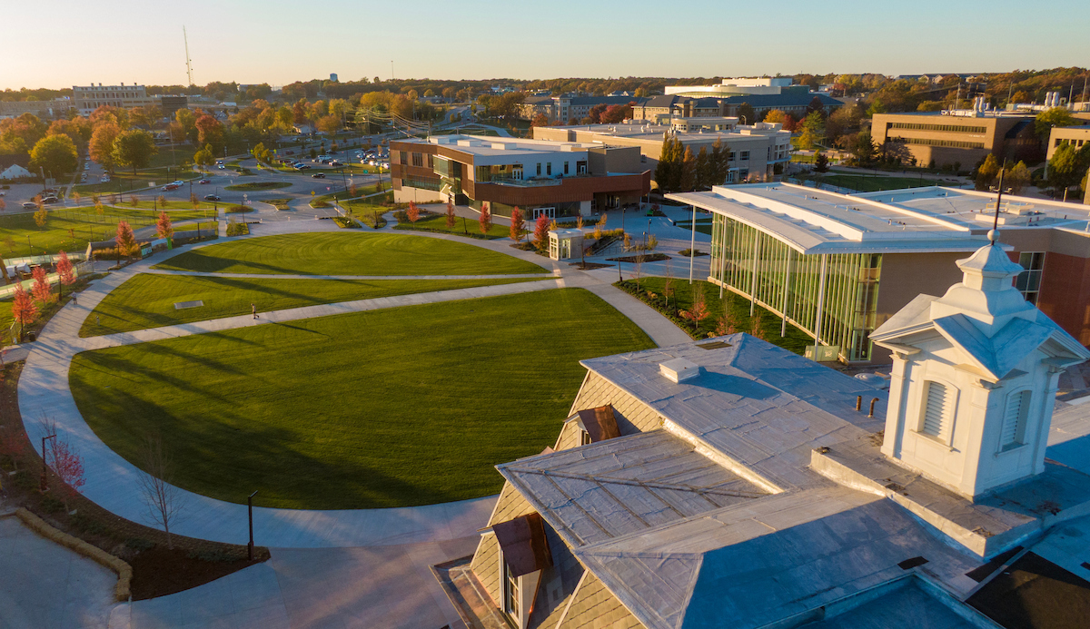 Aerial view of a campus green surrounded by curved walkways and a mix of modern and historic buildings, with autumn trees and additional campus buildings in the background.