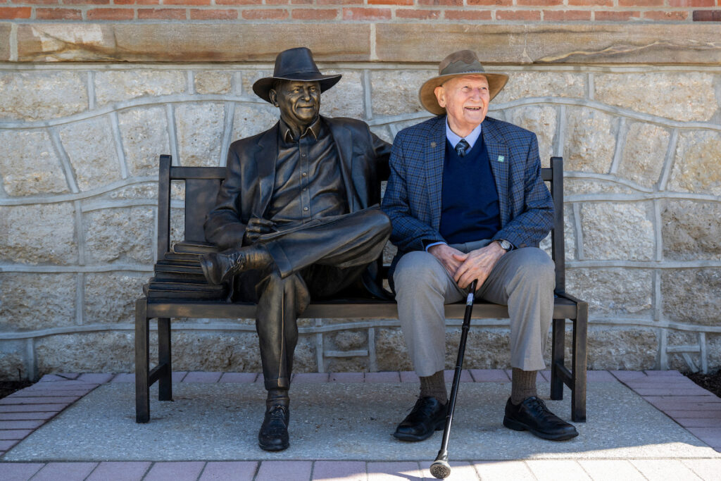 A person in a brimmed hat sits on a bench beside a life-size bronze statue in a similar seated pose, against a stone-and-brick wall.