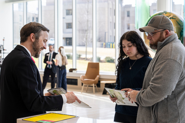 A university representative speaks with two visitors in a bright campus lobby while reviewing informational brochures. Large windows overlook the campus outside, and additional visitors are visible in the background.