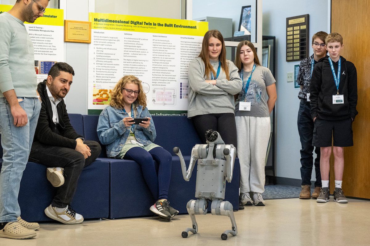 Students and visitors watch a small four-legged robot move across the floor during a poster session. One attendee seated nearby operates a handheld controller, while research posters and display boards are visible in the background.