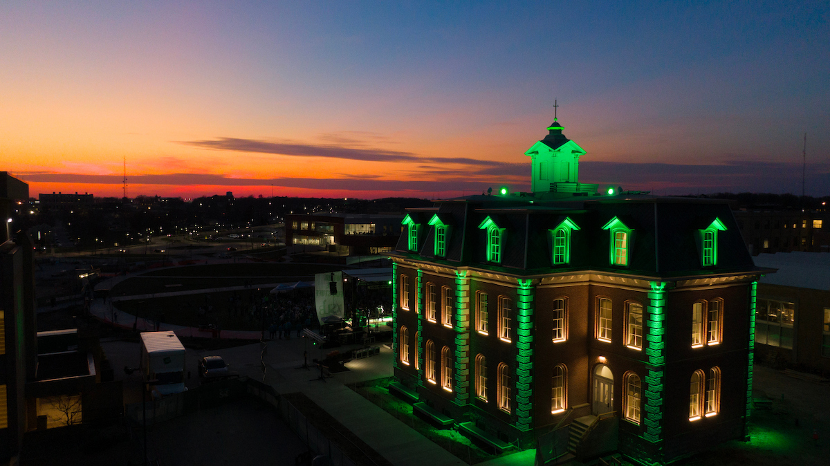 Historic brick building illuminated in green against a blue-and-orange sunset sky, with warm light glowing from the windows.