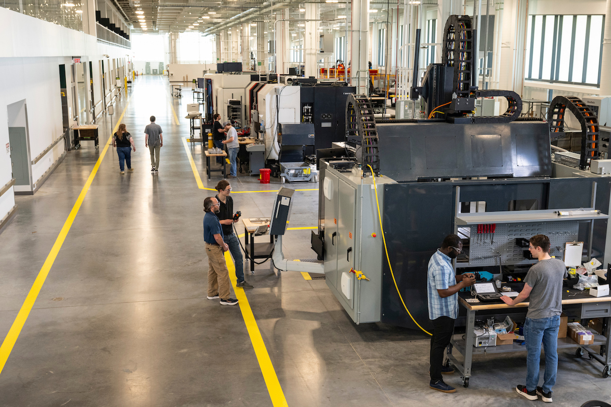 People work at large industrial machines in a spacious manufacturing and research facility, with rows of equipment extending through the building.