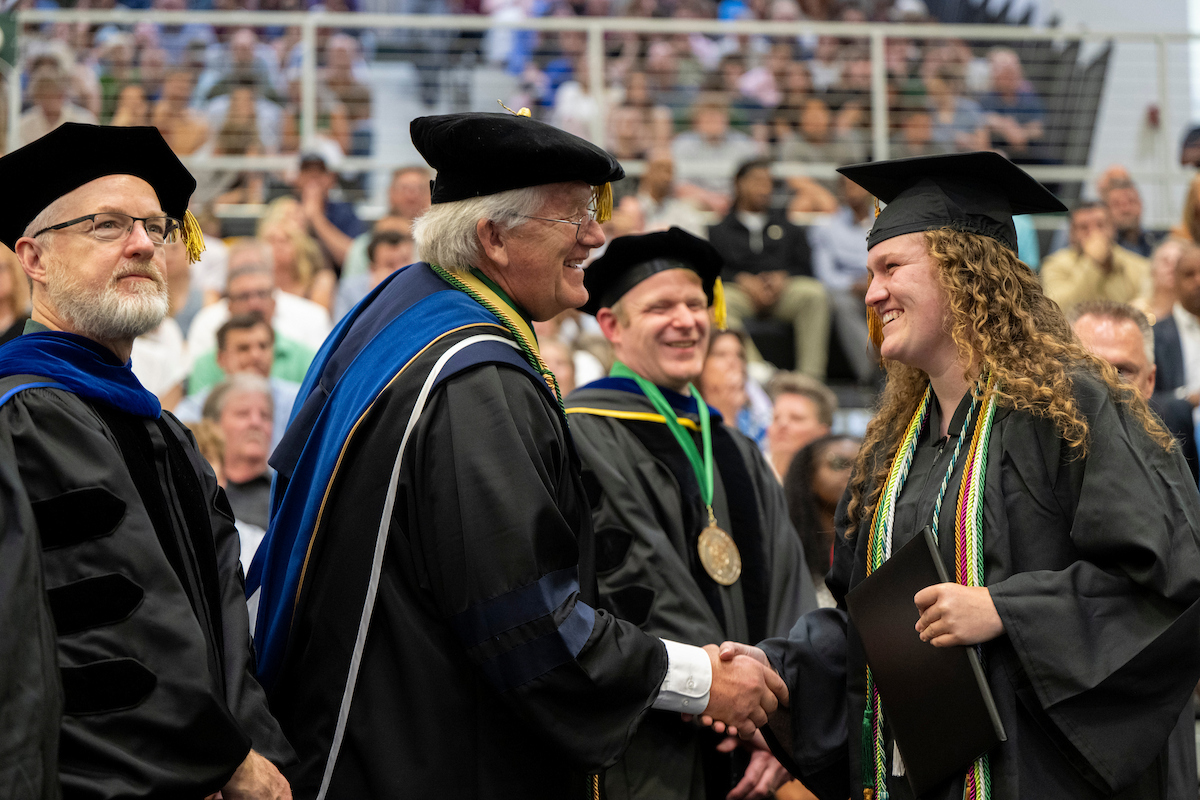 A graduating student in cap and gown shakes hands with a university official during a commencement ceremony while holding a diploma cover. Other regalia-clad officials stand nearby, and audience members fill the background seating area.